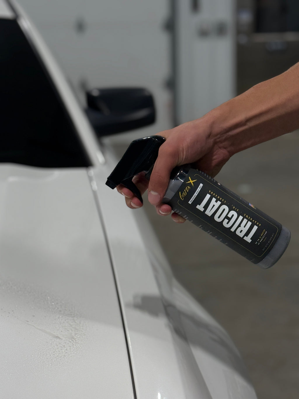 Close-up of a GoldenX Tricoat graphene spray wax bottle held beside the fender of a white car