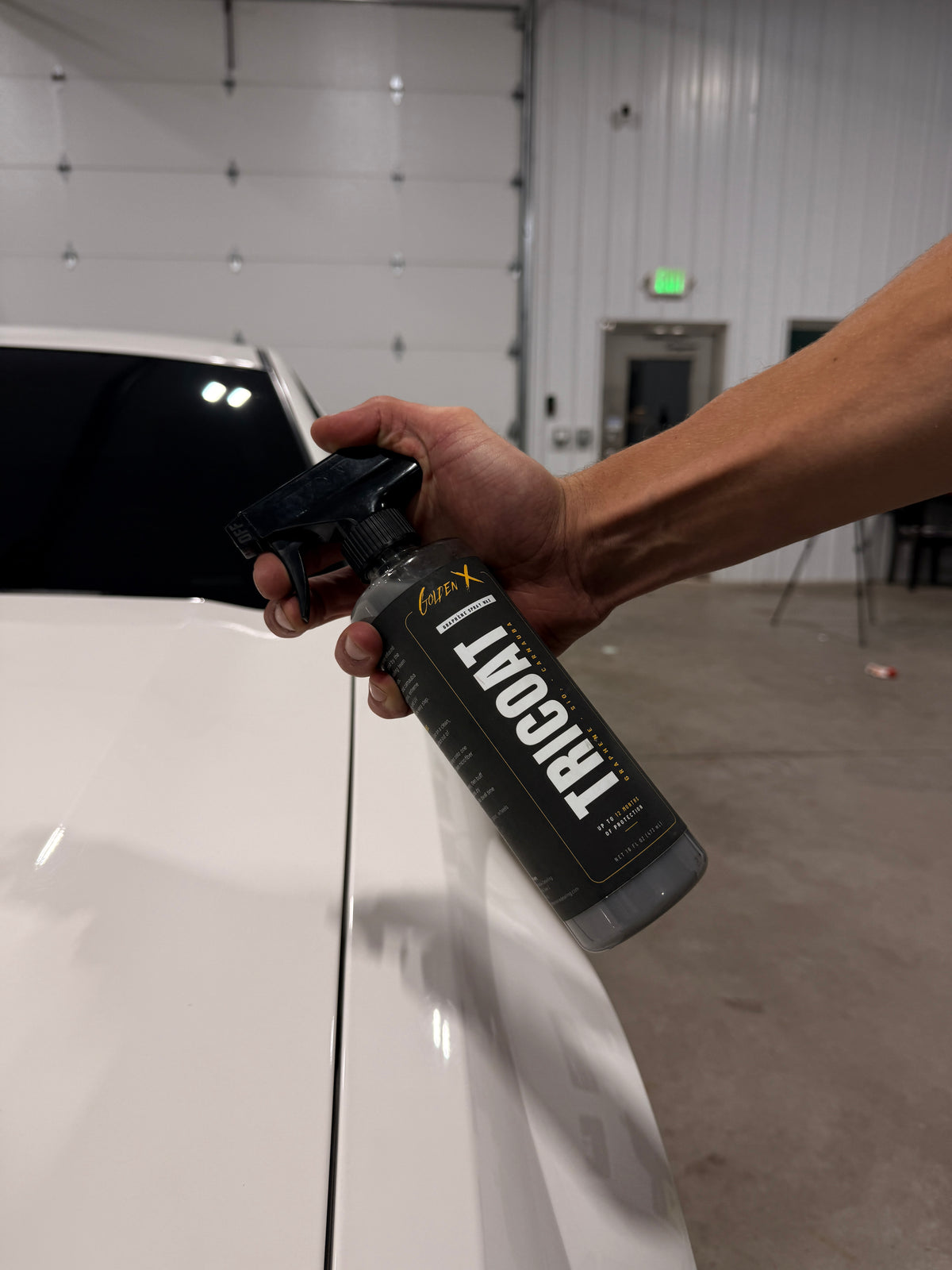 Hand holding a GoldenX Tricoat graphene spray wax bottle over the hood of a freshly detailed white car